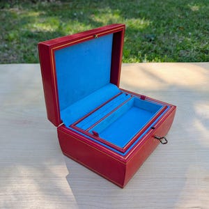 May include: Red jewelry box with a blue velvet interior and gold trim. The box is open, revealing compartments for storage. A small key is in the lock. The box is on a wooden surface.