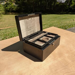May include: An open, black jewelry box with a light-colored interior and multiple compartments. The box has a keyhole and a small key. The box is sitting on a light-colored wooden surface, with a green lawn in the background.