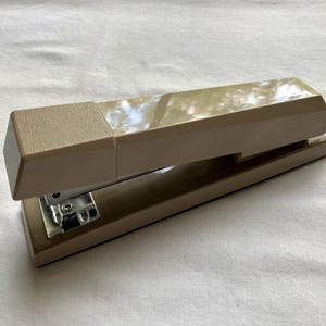 May include: A beige desktop stapler with a glossy finish on the top lever and a textured matte finish on the rear section. The device features a metallic silver internal mechanism visible near the front hinge, designed for fastening paper documents.