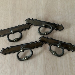 May include: Four antique brass drawer pulls with a decorative, scalloped edge design. Each pull features a rectangular backplate and a round, ring-shaped handle. The hardware is a warm, aged brass color, suggesting vintage or retro style.