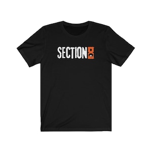 Op de afbeelding: Zwart T-shirt met korte mouwen met de tekst "SECTION 8" in witte letters en een gestileerde oranje "8" afbeelding.
