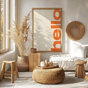 Puede incluir: Un interior de sala de estar con un sofá, dos taburetes, un puf tejido y una impresión enmarcada con la palabra "hello" en letras naranjas. La habitación está decorada con hierba de la pampa seca y una pared colgante tejida.