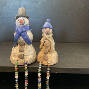May include: Two ceramic snowman figurines with crackled white and tan surfaces. The larger snowman wears a grey top hat and blue scarf, while the smaller one has a blue cap and tan scarf. Both feature dangling legs made of colourful glass beads.