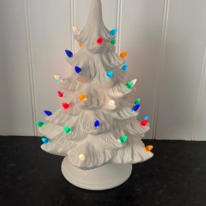 May include: A white ceramic tabletop Christmas tree with a matte finish, featuring a bright red star topper and numerous small, multi-coloured translucent plastic bulbs inserted into the branches, all resting on a circular white base.