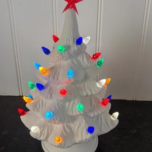May include: A white ceramic tabletop Christmas tree featuring a textured, moulded branch design. The tree is illuminated with small, multi-coloured plastic bulbs in red, blue, green, orange, and clear, topped with a translucent red star.
