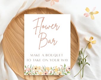 May include: A white sign with the words "Flower Bar" in a cursive font. The sign also reads "Make a bouquet to take on your way" and features a floral border with yellow and pink flowers. The sign is on a wooden plate.