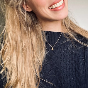 May include: A gold-colored necklace with a small, round pendant hangs against a dark blue cable-knit sweater. The necklace is delicate and simple. The person wearing the necklace has long blonde hair and is smiling.