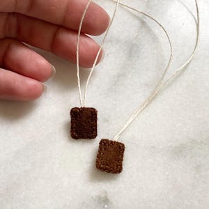 May include: Two small brown felt squares with stitched edges are attached to white string. The string is looped and held by a hand.
