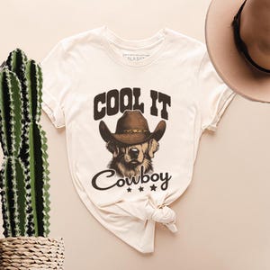 May include: A white t-shirt with a brown and black illustration of a dog wearing a cowboy hat. The text "Cool It Cowboy" is printed on the shirt.