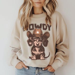May include: A beige sweatshirt with a brown dachshund wearing a cowboy hat and the word "COWDY" printed on the front.