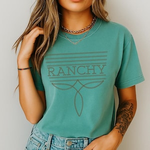 May include: Teal-colored t-shirt with the word "RANCHY" in a brown, stylized font. The design includes horizontal lines above the word and a decorative shape below. The shirt has short sleeves and a crew neck.