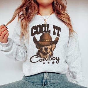 May include: A gray sweatshirt with the text "Cool It Cowboy" and a cartoon image of a golden retriever wearing a brown cowboy hat.
