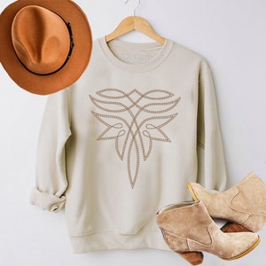May include: A cream-colored sweatshirt with a brown embroidered design on the front. The design is a stylized geometric pattern. The sweatshirt is hanging on a wooden hanger. A brown hat and brown ankle boots are also pictured.