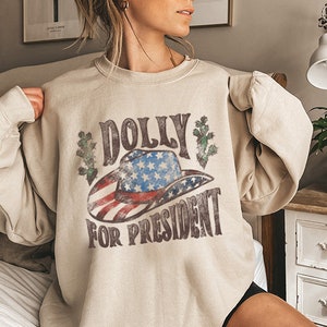 May include: A beige sweatshirt with a vintage-style graphic print of a cowboy hat with an American flag design and the text "DOLLY FOR PRESIDENT".