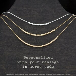 May include: Three gold-filled or sterling silver necklaces with beads in the shape of dots and dashes, arranged in a layered fashion on a dark brown surface. The necklaces are personalized with a message in Morse code. The text below the necklaces reads "Personalized with your message in morse code".