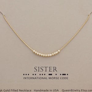 May include: A delicate gold-filled necklace with a series of small beads and cylindrical shapes, spelling out "SISTER" in Morse code. The necklace is presented on a neutral-toned card with the words "SISTER" and "INTERNATIONAL MORSE CODE" printed below.