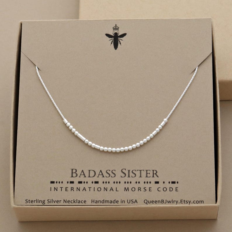 Sterling silver morse code necklace in a box. The dainty necklace has sterling silver beads that spells out the words 'Badass Sister' in morse code. The box is brown with QueenBJwlry bee logo and the words 'Sterling Silver Necklace Handmade in USA QueenBJwlry.Etsy.com'.
