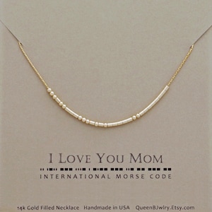 May include: A gold-filled necklace with a Morse code design. The necklace features a delicate gold chain and a series of small, cylindrical beads and round beads. The card reads "I Love You Mom" and "International Morse Code".
