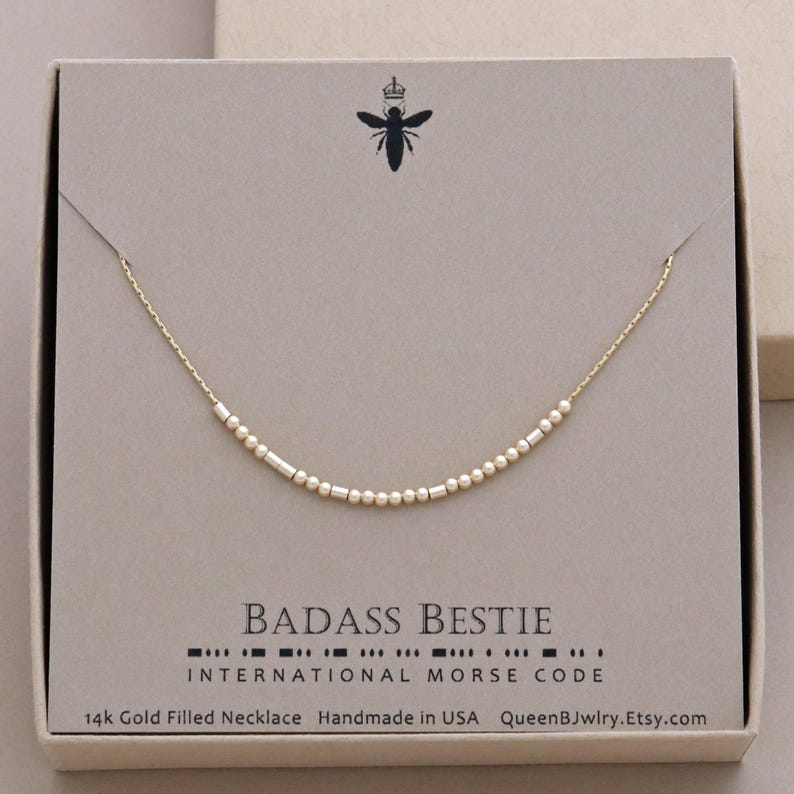 Best Friend necklace with a chain and small gold beads. The beads spell out the words 'Badass Bestie' in international morse code. The text below the necklace reads 'INTERNATIONAL MORSE CODE', '14k Gold Filled Necklace', 'Handmade in USA', and 'QueenBJwlry.Etsy.com'.