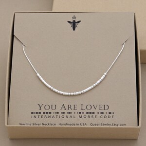 A Sterling silver morse code necklace in a box. The custom necklace has sterling silver beads that spells out the words 'You are Loved' in morse code. The gift box is kraft brown with QueenBJwlry bee logo and the words 'Sterling Silver Necklace Handmade in USA QueenBJwlry.Etsy.com'.