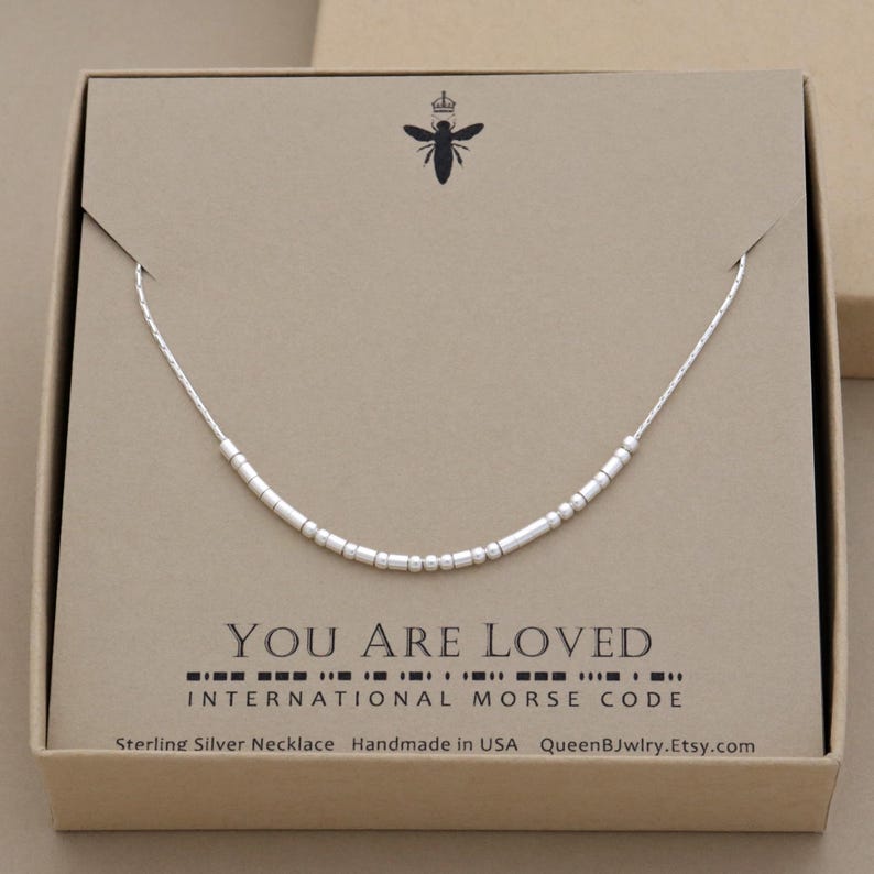 A Sterling silver morse code necklace in a box. The custom necklace has sterling silver beads that spells out the words 'You are Loved' in morse code. The gift box is kraft brown with QueenBJwlry bee logo and the words 'Sterling Silver Necklace Handmade in USA QueenBJwlry.Etsy.com'.