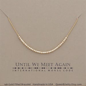 May include: A delicate gold-filled necklace with a Morse code design. The necklace features a series of small, cylindrical and spherical beads arranged on a fine chain. The card reads "UNTIL WE MEET AGAIN" in Morse code. The text "INTERNATIONAL MORSE CODE" is also present.