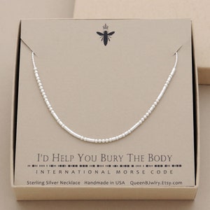 A sterling silver necklace in a box. The necklace is made of sterling silver beads that spell out the words 'I'D HELP YOU BURY THE BODY' in morse code. 'INTERNATIONAL MORSE CODE' 'Sterling Silver Necklace Handmade in USA QueenBJwlry.Etsy.com'.