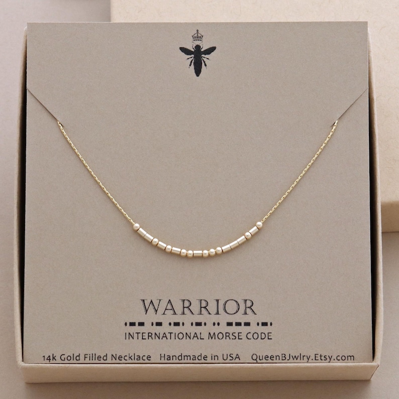 Dainty gold necklace with a chain and small gold beads. The beads spell out the word 'WARRIOR' in morse code. The text below the necklace reads 'INTERNATIONAL MORSE CODE', '14k Gold Filled Necklace', 'Handmade in USA', and 'QueenBJwlry.Etsy.com'.
