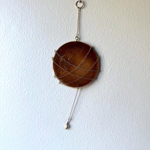 May include: A round, brown wall hanging with a metallic gold chain design. The circular piece is suspended from a small ring and chain, with a dangling bell-shaped charm. The wall decor is mounted on a white wall.