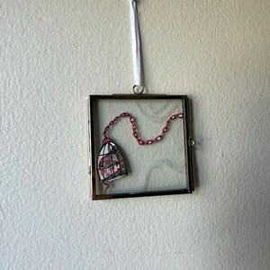 May include: A square, framed artwork featuring a hand-drawn illustration of a cage with a pink chain. The cage contains a sketched face with pink hair. The artwork is suspended by a white ribbon.