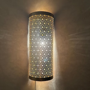 Könnte beinhalten: Zylindrische Wandleuchte mit perforiertem Metallschirm. Der Schirm ist dunkelgrau und strahlt ein warmes, diffuses Licht durch die kleinen, gleichmäßig verteilten Löcher aus. Die Leuchte ist an einer strukturierten, hellgrauen Wand befestigt.