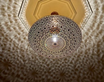 May include: A round, gold-toned metal pendant light fixture with a intricate, geometric pattern. The light fixture is hanging from a ceiling and casting a patterned shadow on the wall.