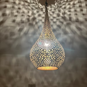 May include: A white, teardrop-shaped pendant light with intricate cut-out patterns. The light fixture is suspended from a chain and casts a patterned glow on the surrounding surface. The interior glows with a warm light.