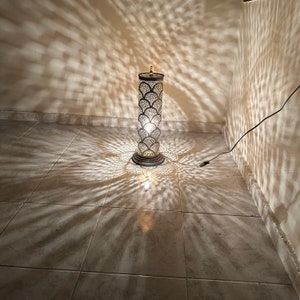 May include: A tall, silver floor lamp with a decorative, patterned metal shade. The lamp casts intricate shadows on the tiled floor.