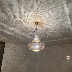 May include: A decorative pendant light fixture with a conical shape, featuring a perforated metal shade. The light casts intricate patterns on the ceiling and walls. The fixture has a gold-colored top and a warm, inviting glow.