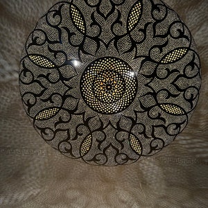 May include: A round, black metal lampshade with a decorative, intricate pattern of cut-outs. The pattern features a central circle with a smaller circle inside, surrounded by swirling designs.