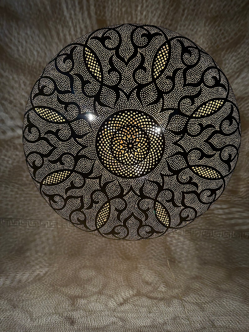 May include: A round, black metal lampshade with a decorative, intricate pattern of cut-outs. The pattern features a central circle with a smaller circle inside, surrounded by swirling designs.