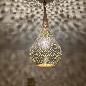 May include: A decorative, hanging pendant light with a teardrop shape. The light fixture is made of a metal material with intricate cut-out patterns. The light casts a patterned shadow on the surrounding wall. The light is suspended from a chain.