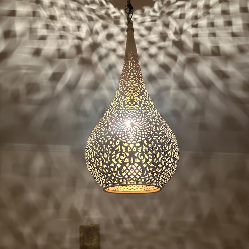 May include: A decorative, hanging pendant light with a teardrop shape. The light fixture is made of a metal material with intricate cut-out patterns. The light casts a patterned shadow on the surrounding wall. The light is suspended from a chain.