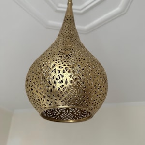 May include: A gold-colored, teardrop-shaped pendant light fixture. The metal shade features intricate cut-out patterns, allowing light to filter through. The fixture is suspended from the ceiling, showcasing its decorative design and warm metallic finish.