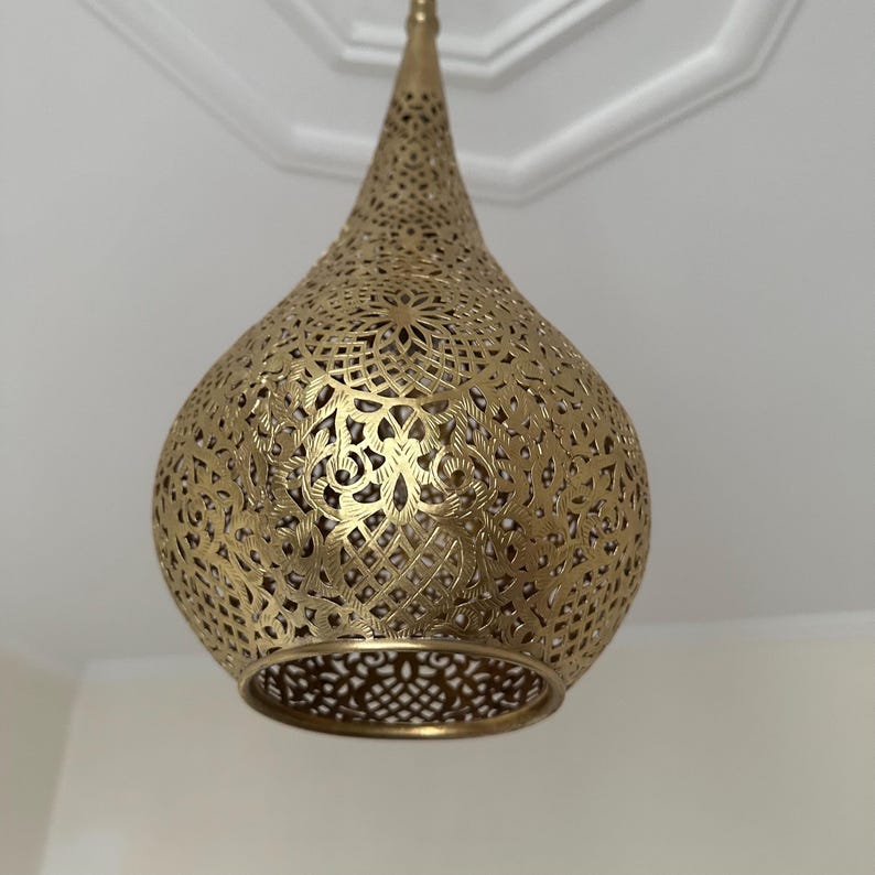 May include: A gold-colored, teardrop-shaped pendant light fixture. The metal shade features intricate cut-out patterns, allowing light to filter through. The fixture is suspended from the ceiling, showcasing its decorative design and warm metallic finish.