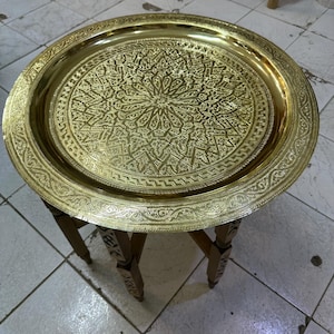 Pode incluir: Mesa de bandeja redonda de latão dourado com um intrincado padrão geométrico em relevo no tampo. A mesa tem uma base de madeira com pernas decorativas. Uma peça decorativa, com cerca de 60 cm de diâmetro.