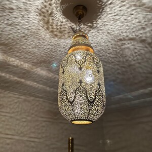 May include: A gold metal pendant light fixture with a decorative, intricate, cut-out design. The light fixture is hanging from a chain.