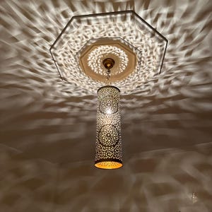 May include: A gold metal pendant light fixture with a cylindrical shade featuring intricate cut-out designs. The light casts a patterned shadow on the ceiling.