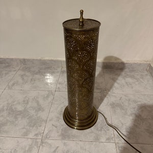 May include: A gold metal floor lamp with a cylindrical shade featuring a decorative, intricate pattern of cut-outs. The lamp has a round base with a decorative trim.