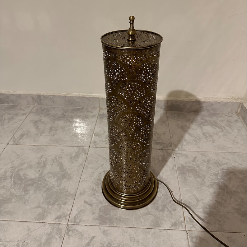 May include: A gold metal floor lamp with a cylindrical shade featuring a decorative, intricate pattern of cut-outs. The lamp has a round base with a decorative trim.