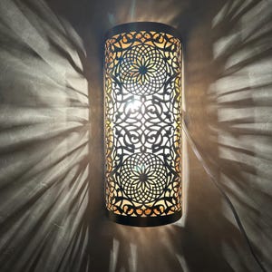 Pode incluir: Uma caneca de cerâmica branca com uma alça preta e o texto "But First, Coffee" em negrito preto. A caneca está sobre um fundo desfocado, sugerindo um ambiente acolhedor.