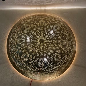 Op de afbeelding: Een ronde, gouden metalen plafondlamp met een decoratief, ingewikkeld, gepatroneerd ontwerp. De lamp is verlicht en geeft een warm licht.