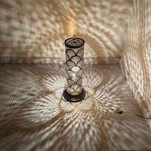 May include: A tall, cylindrical floor lamp with a decorative metal shade featuring a geometric pattern. The lamp is lit, casting intricate shadows on the tiled floor.