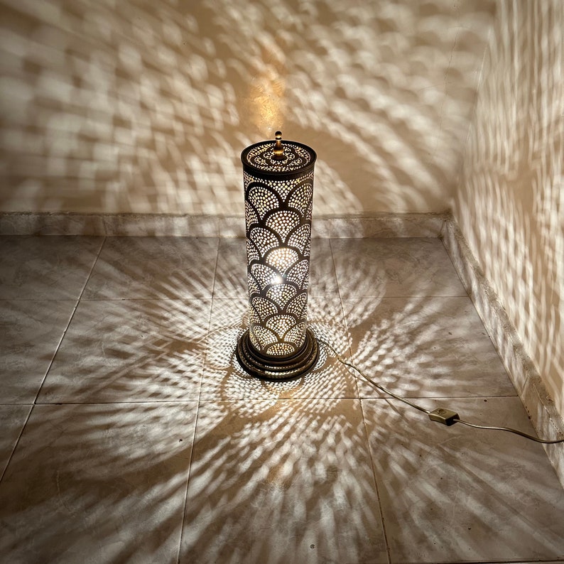 May include: A tall, cylindrical floor lamp with a decorative metal shade featuring a geometric pattern. The lamp is lit, casting intricate shadows on the tiled floor.